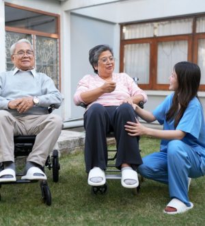 Two seniors are sitting in a garden. One is in a wheelchair. A caregiver is talking to them and offering support. The setting is bright with grass and windows visible behind them.